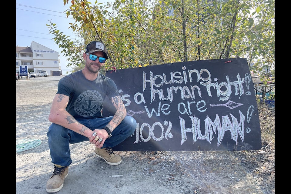 Craig Strachan, who helped organize the tent encampment by Fraser Tower, poses beside a sign in front of the site. The sign reads: "Housing is a human right! We are 100 per cent human."