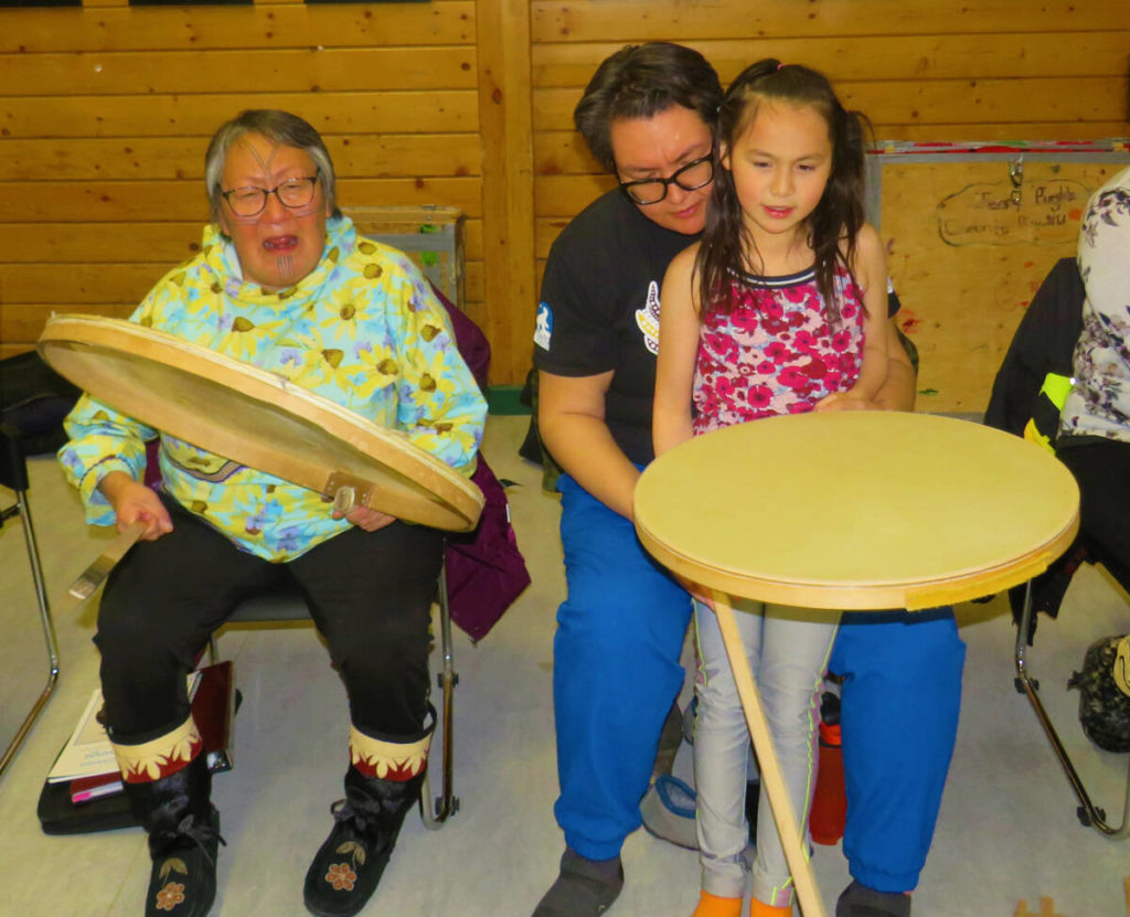 ᐱᔭᕆᐅᖅᓴᓂᕐᒥ ᕿᓚᐅᔾᔭᕐᓂᕐᒥ ᐊᑕᐅᓯᐅᓪᓗᓂ ᐊᔪᙱᓐᓂᐅᔪᒥ ᐃᓕᓴᕋᐃᓐᓂᐅᔪᒥ ᕼᐅᖅᑯᓪᓛᖅᑎᓪᓗᑎᑦ ᐱᕼᐃᓂᑦ ᓯᕗᕚᑦᑎᓐᓂ. ᐅᓇ ᐃᓄᑐᖃᖅ / ᐃᓕᓴᐃᔨ ᔪᓕᐊ ᕼᐊᐅᒐᒃ ᐅᔩᓇ ᐃᙱᖅᑐᖅ ᐱᓯᕐᓂ ᐅᓪᓘᔪᓄᑦ ᐊᖑᓇᓱᖕᓂᕐᒥ ᐊᒻᒪ ᓄᓇᒥ ᐆᒪᓇᓱᐊᕐᓂᕐᒥ. ᐃᒃᓯᕚᖅᑐᖅ ᕼᐊᐅᒐᖕᒥ ᓂᑰᓪ ᐅᐃᓪᑳᒃᔅ–ᖑᕗᖅ ᐊᒻᒪ ᐅᔪᕈᖓ, 8–ᓂ–ᐅᑭᐅᓕᒃ ᐊᕝᕙ ᕕᓕᐳᕋᓐ–ᐅᐃᓪᑳᒃᔅ. ᐊᕝᕙ ᐃᓕᑦᑎᓇᓱᐊᕈᒪᓚᐅᖅᐳᖅ ᕿᓚᐅᔾᔭᕐᓂᕐᒥ ᒪᒃᑯᒃᑎᓪᓗᒍ. ᐱᔪᒪᕐᔪᐊᖅᑐᖅ. ᐱᒻᒪᕆᐅᓂᖅᓴᐅᕗᖅ ᐊᖓᔪᖅᑳᑦ ᐊᓈᓇᑦᑎᐊᑦ ᐊᑖᑕᑦᑎᐊᑦ ᓱᕈᓯᕐᒥ ᐃᓚᐅᖃᑕᐅᑎᓪᓗᒋᑦ ᐱᔭᕆᐅᖅᓴᑎᓪᓗᒋᑦ .Practising how to beat the drum is one skill to easily learn as you huqqullaaq pihiit of our ancestors. Here is Elder/teacher Julia Haogak Ogina singing the songs of the days of hunting and living off the land. Sitting beside Haogak is Nicole Wilcox and her niece, eight-year-old Avva Villebrun-Wilcox. Avva wanted to try to learn how to beat the drum at a young age. So determined. It is more special when the parents and grandparents of a child come with them to the practices. Navalik Tologanak/NNSL photo