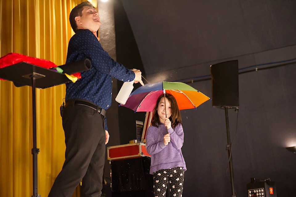 Charlotte Mayer keeps the “rain” away while assisting Leif David during his magic show at the Salmar Classic on Saturday, Jan. 20, 2024. The show was the first of two Saturday Unplug and Play performances by David for Family Literacy Week, organized by the Literacy Alliance of the Shuswap Society. (Lachlan Labere-Salmon Arm Observer) 