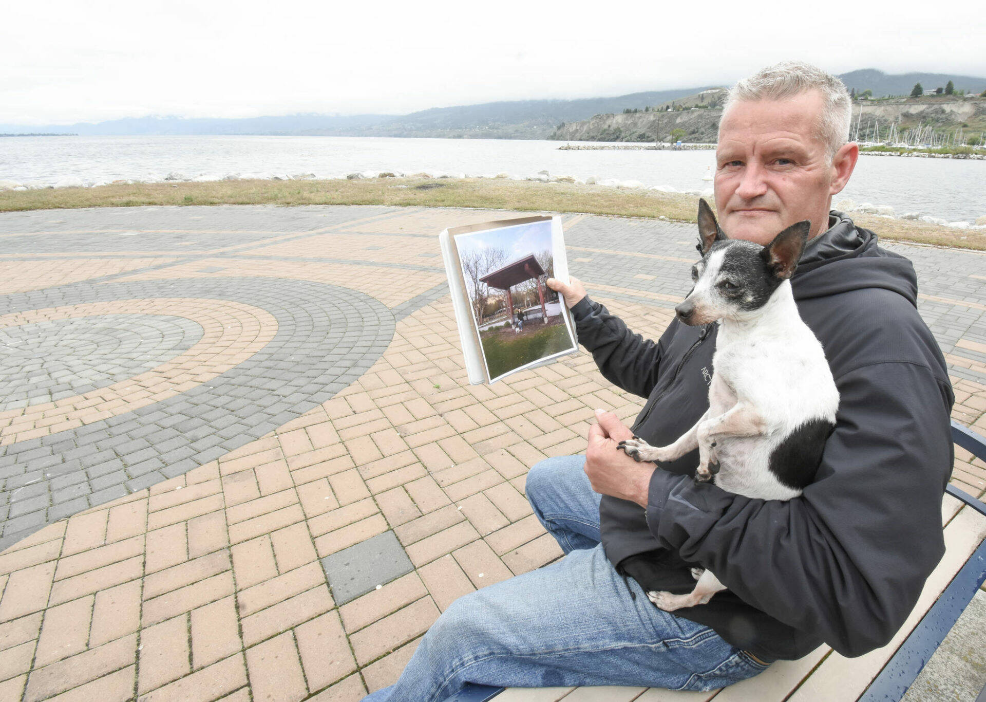 Gord Portman and his dog Zippy sit at Marina Way Park while holding a picture of a bench he would like to have installed in memory of those who have lost their lives to an overdose. (Mark Brett photo)