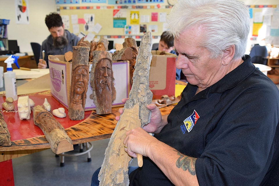 Métis carver and elder John Sayer works at the Storefront School in Salmon Arm on Thursday, Nov. 19, helping students learn to carve and sharing his culture, while teacher Robin Wiens and student William Dicer try their hand at carving. (Martha Wickett - Salmon Arm Observer)