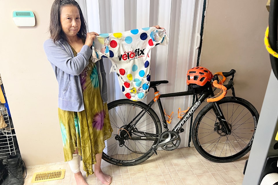 Yuriko Marshall holds up the torn jersey she was wearing when she crashed after a close pass by a driver in a pickup pulling a trailer. She stands next to her brightly coloured and broken helmet and bicycle handlebars.